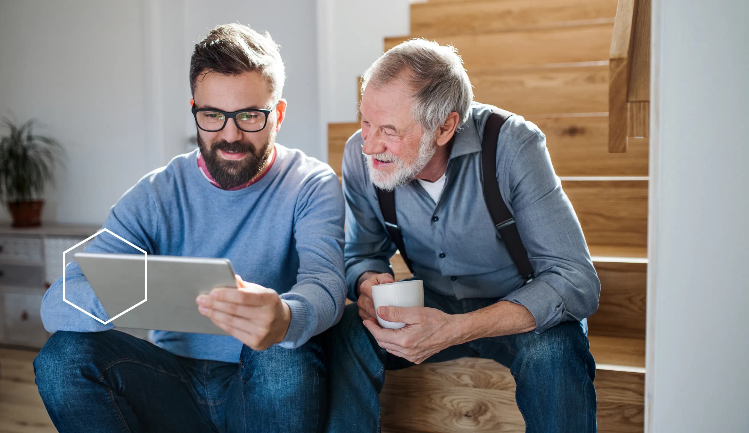 A son holding a tablet and his father looks at the screen smiling, while holding a coffee cup.