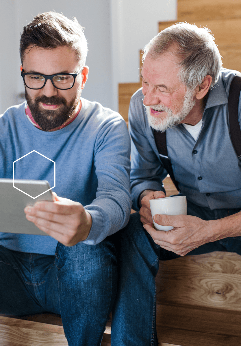 A son holding a tablet and his father looks at the screen smiling, while holding a coffee cup.