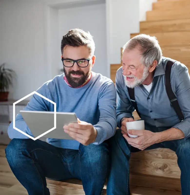 A son holding a tablet and his father looks at the screen smiling, while holding a coffee cup.